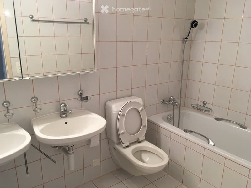 Bathroom with white tiled walls and floor, featuring a toilet, sink, bathtub, and mirror. The sink has a chrome faucet, and the bathtub has a handheld showerhead.
