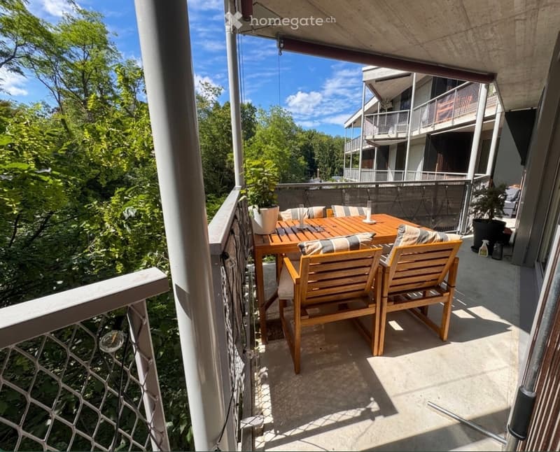 Balcony with wooden table and chairs, overlooking a green, tree-filled area.