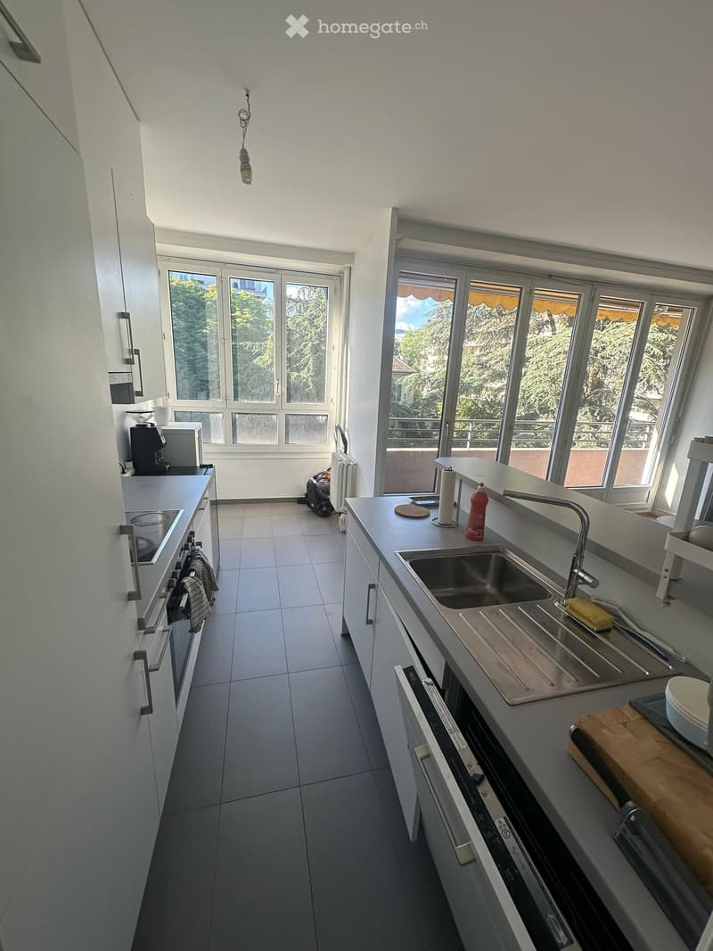 Modern kitchen with white cabinets, stainless steel sink, oven, coffee maker, and various small appliances. Large windows provide natural light and a view of greenery.