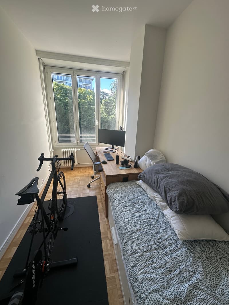 A small, furnished bedroom with a single bed, desk, chair, computer, and bicycle. The room has wooden flooring and large windows with a view of trees.