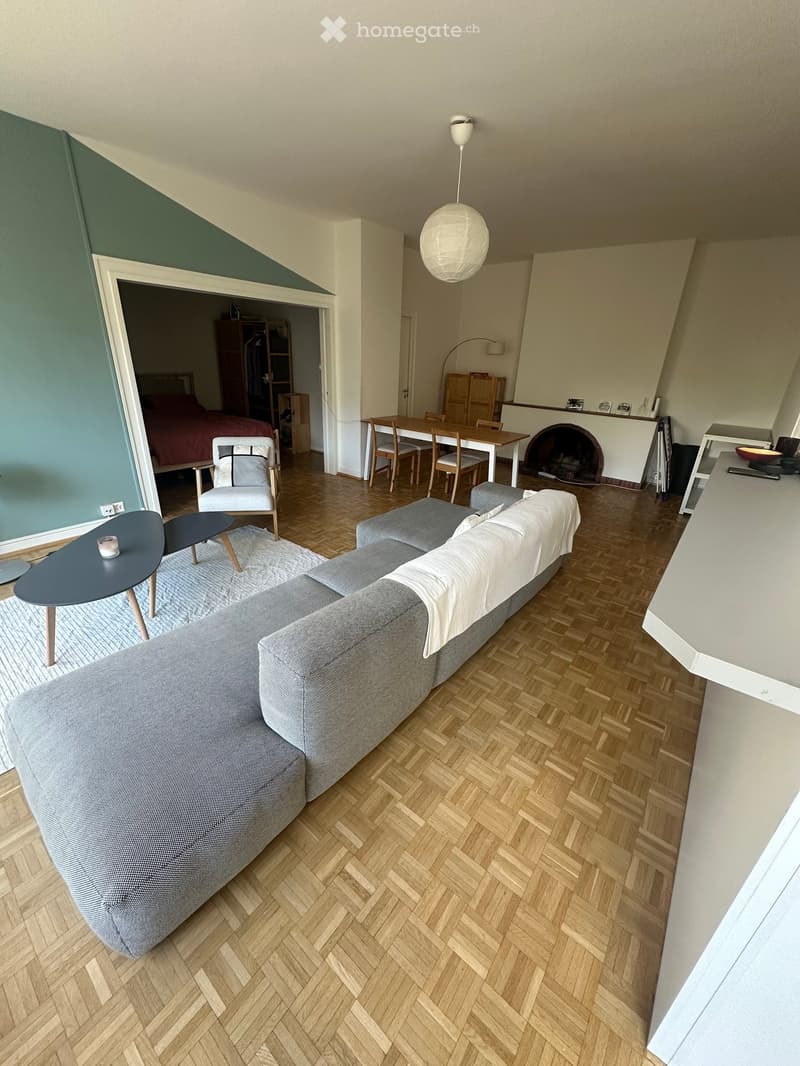 Modern living room with grey sectional sofa, coffee table, dining area, and adjacent bedroom visible through an open doorway.