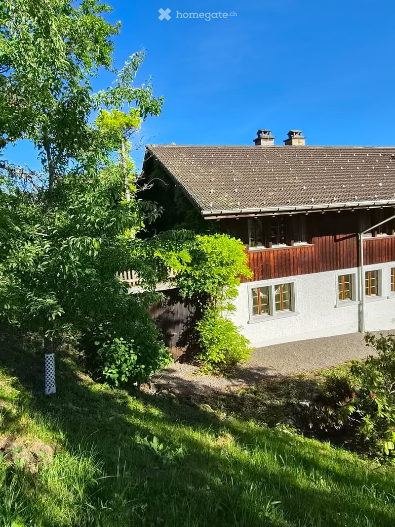 A two-story house with a tiled roof, white walls, and wooden trim. The house features multiple windows and a small balcony. The surrounding area includes greenery and a gravel driveway.