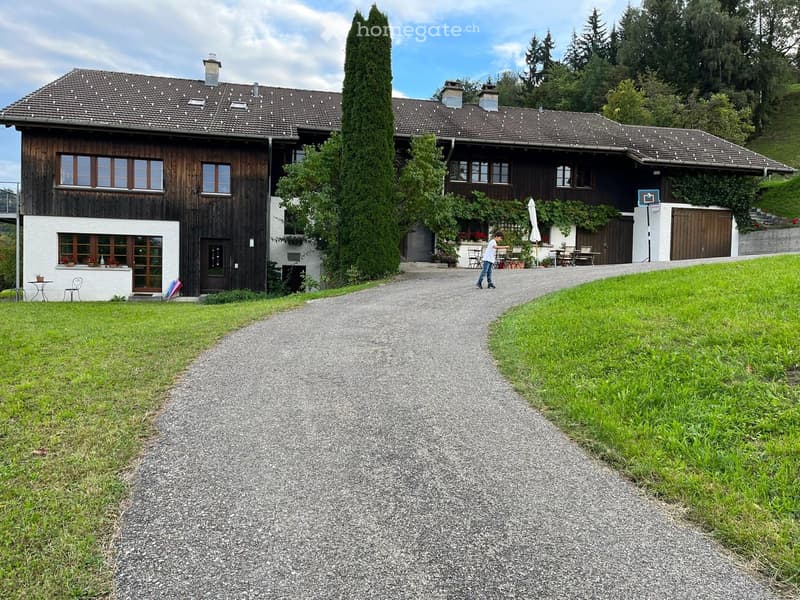 A two-story wooden house with a tiled roof, multiple windows, and a covered outdoor seating area. The house is surrounded by greenery and a gravel driveway.
