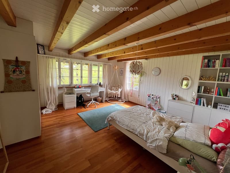 Cozy bedroom with wooden floors, slanted wooden ceiling beams, large windows with white curtains, a bed with white bedding, a desk with a chair, a wicker hanging chair, a bookshelf, and a round mirror on the wall.