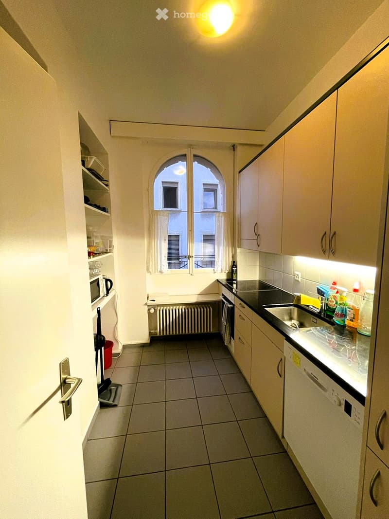 Modern kitchen with white cabinets, tiled floor, and a window with white curtains. Includes a microwave, fridge, sink, dishwasher, and cleaning supplies on the counter.