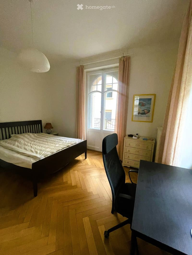 Cozy bedroom with wooden flooring, a double bed with white bedding, a desk with a chair, a small lamp, a dresser, and a framed picture on the wall. The room has large windows with curtains and a view of the outside.