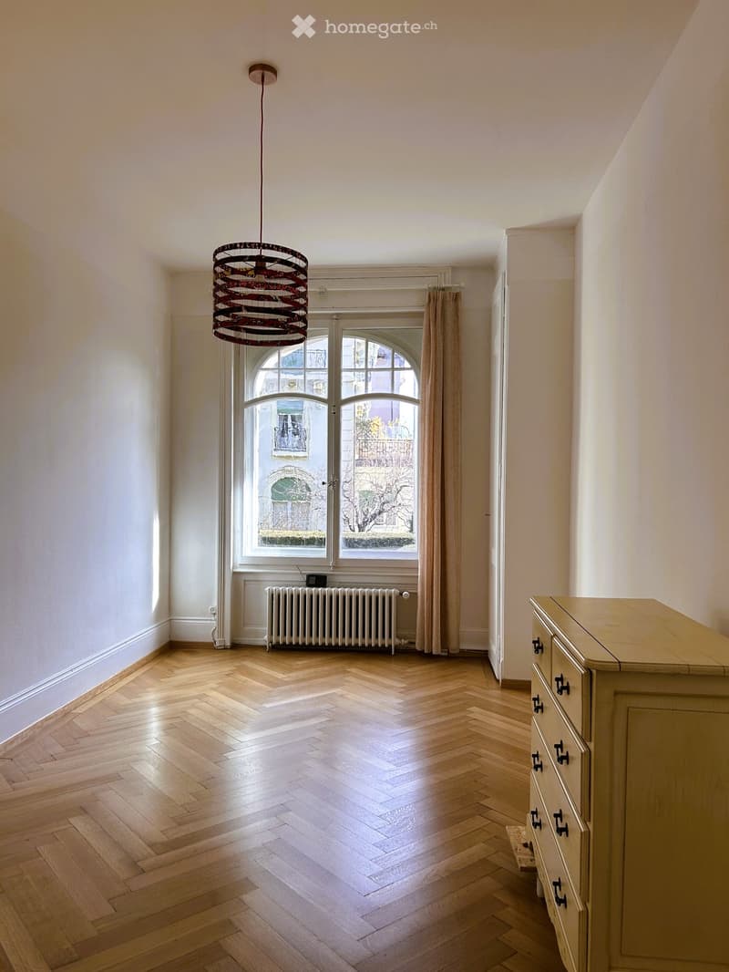 Empty room with wooden herringbone flooring, white walls, large arched windows with curtains, a radiator, and a decorative pendant light fixture.