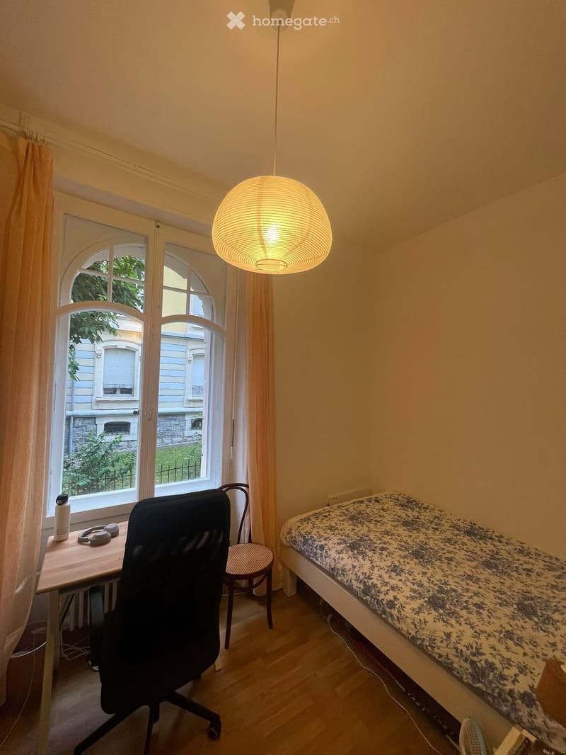 A small, cozy bedroom with a bed, a desk with a chair, a hanging lamp, and a window with curtains.