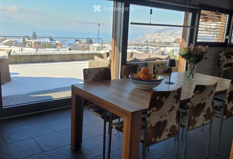 Dining area with a wooden table, chairs with cowhide upholstery, a bowl of oranges, a vase with flowers, and a lamp, overlooking a scenic view of a town and mountains.