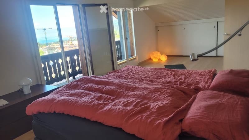 Bright bedroom with large windows offering a scenic ocean view, featuring a neatly made bed with red bedding, a nightstand with a lamp, and decorative ball lamps on the floor.