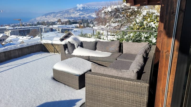 A snow-covered balcony with a modern wicker sofa and chair, overlooking a scenic snowy town and mountains.