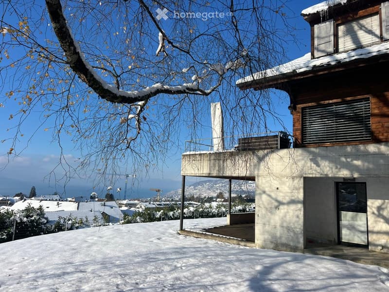 Modern two-story house with a raised ground floor, snow-covered roof, and a balcony overlooking a snowy landscape.