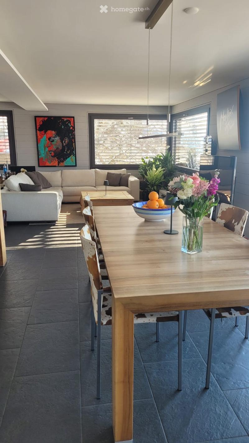 Modern living and dining area with a grey sofa, coffee table, dining table with chairs, vase with flowers, bowl of fruit, and artwork on the wall.