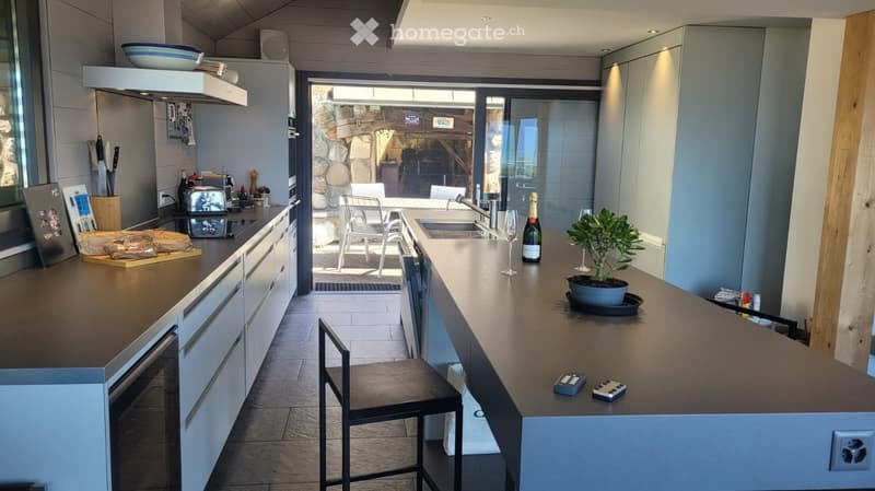 Modern kitchen with white cabinets, dark countertops, and a central island. Features include a sink, a bottle of wine, a potted plant, and a view to an outdoor patio with chairs.