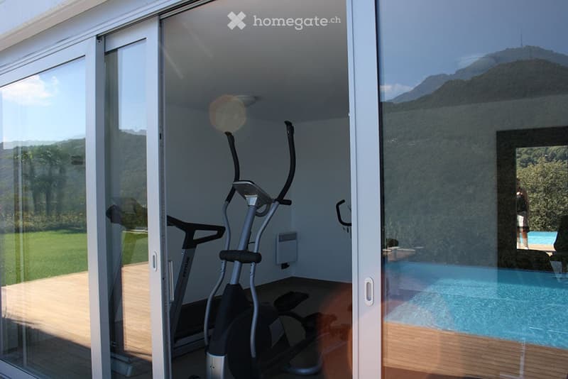 Modern indoor space with large glass doors opening to a wooden deck, featuring an exercise bike and a view of a swimming pool and green landscape.
