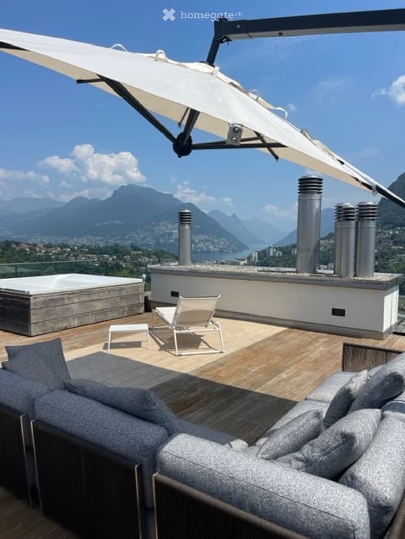 A modern rooftop terrace with a panoramic mountain view, featuring a large retractable umbrella, a sofa, two lounge chairs, and a small table.