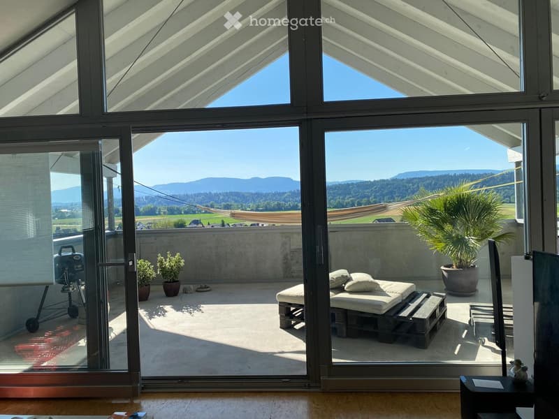 Modern balcony with large glass doors, daybed, stool, potted plants, and a grill, overlooking a scenic mountain landscape.