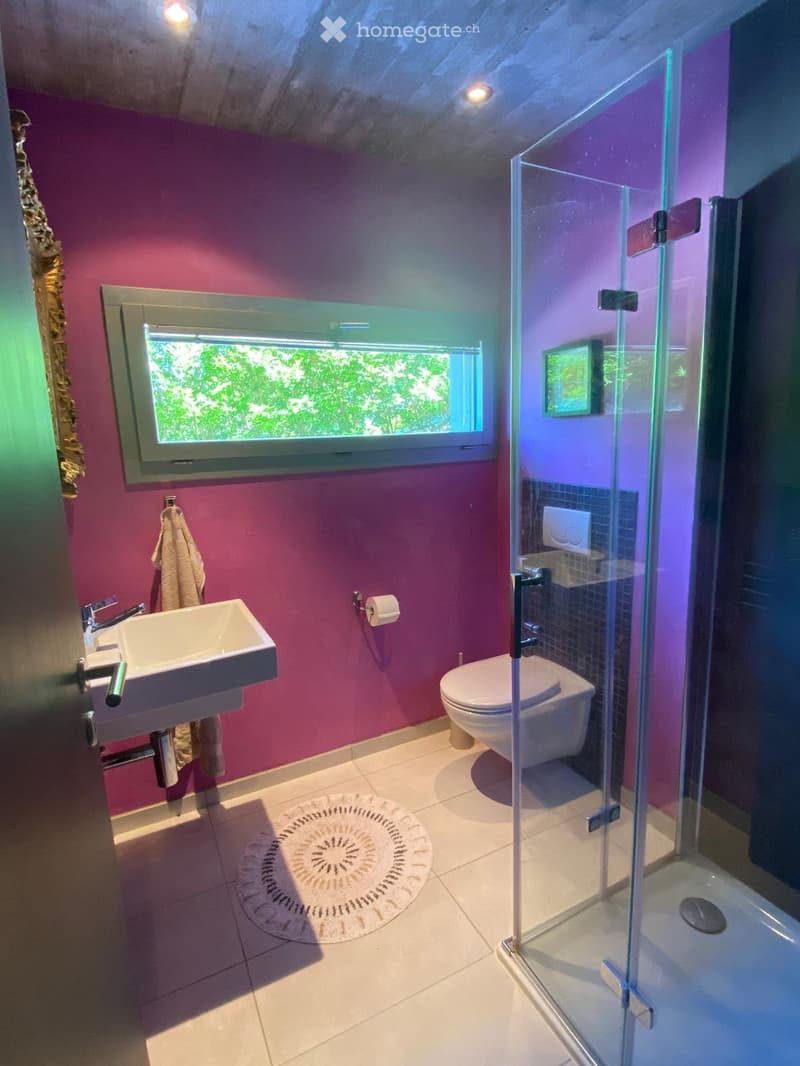Modern bathroom with pink walls, white tiled floor, a sink, toilet, and a glass-enclosed shower. A large window provides natural light, and a towel hangs on the wall.