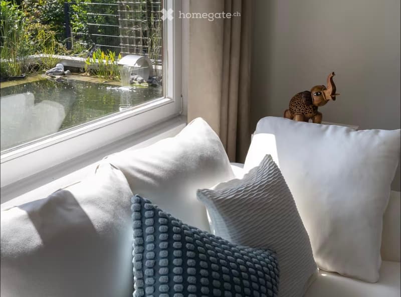A cozy living room corner with a white sofa, multiple pillows, a blue blanket, and a decorative elephant statue on a windowsill, overlooking a serene garden with a pond and water fountain.