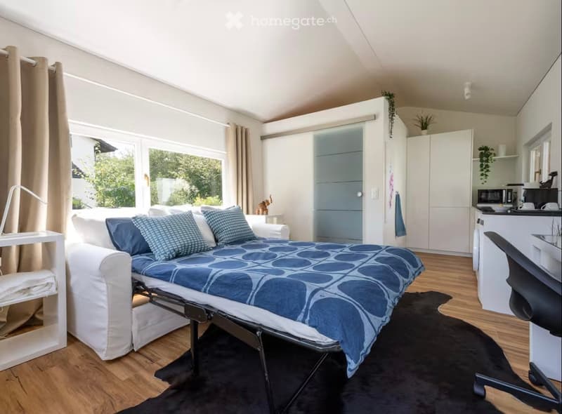 A modern, compact studio with a sofa bed, a small table, a chair, a microwave, a fridge, and a lamp. The room features large windows with curtains and a sleek, minimalist design.