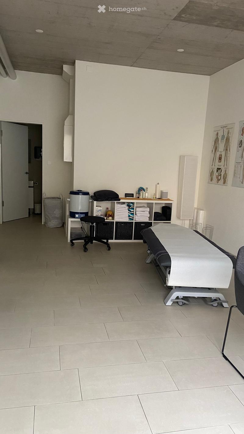 A clean, well-lit treatment room with a tiled floor, a treatment table, a cabinet with towels and supplies, a small refrigerator, and a chair.