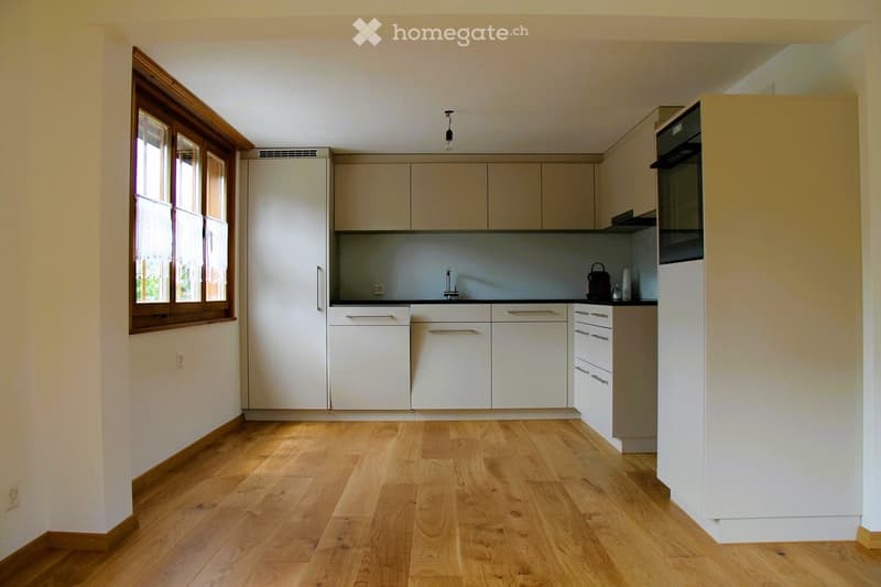 Modern kitchen with white cabinets, wooden flooring, and a large window. Includes an oven, a faucet, and a teapot on the counter.