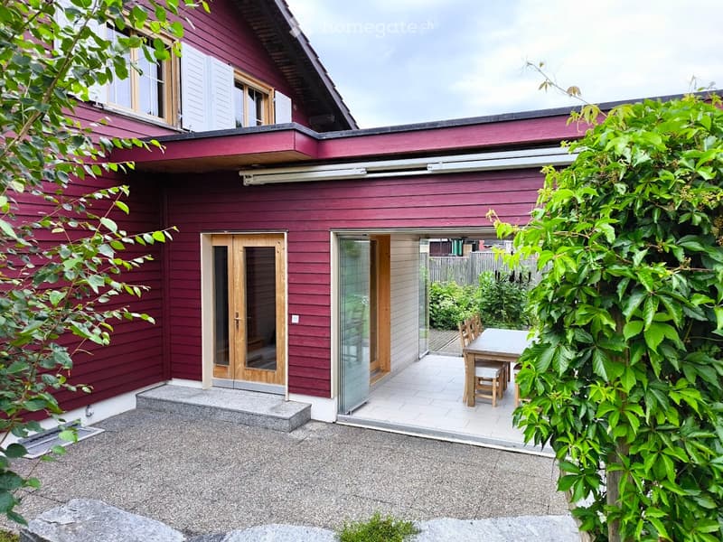 Two-story house with red wooden exterior, white-framed windows, and a small patio area with a table and chairs.