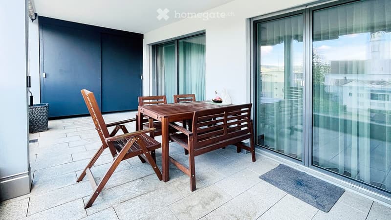 Balcony with tiled floor, wooden table, chairs, and bench, large glass windows, and a view of the city.