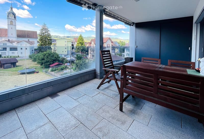 Balcony with tiled floor, wooden table and chairs, large windows offering a view of the town and surrounding landscape.