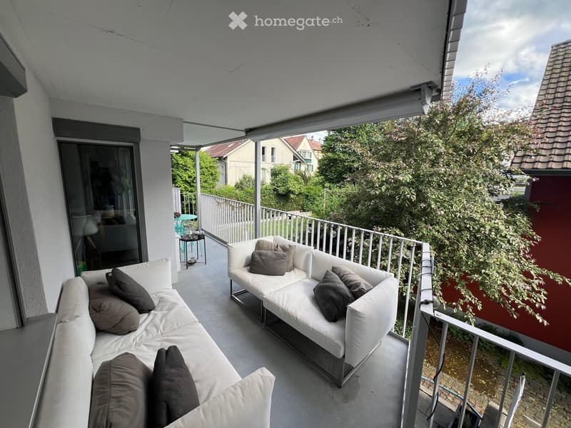 Balcony with white sofa and cushions, overlooking a green area with trees and neighboring houses.