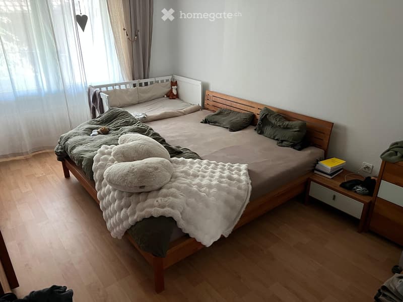 Cozy bedroom with wooden flooring, a large bed with green and white bedding, pillows, a nightstand with books, and a small decorative item on the wall.
