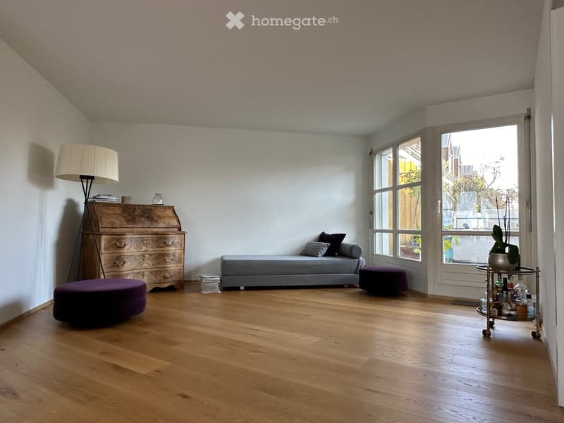 A modern living room with wooden flooring, a grey sofa, a purple ottoman, a wooden chest of drawers, a floor lamp, and a small cart with bottles and a vase.