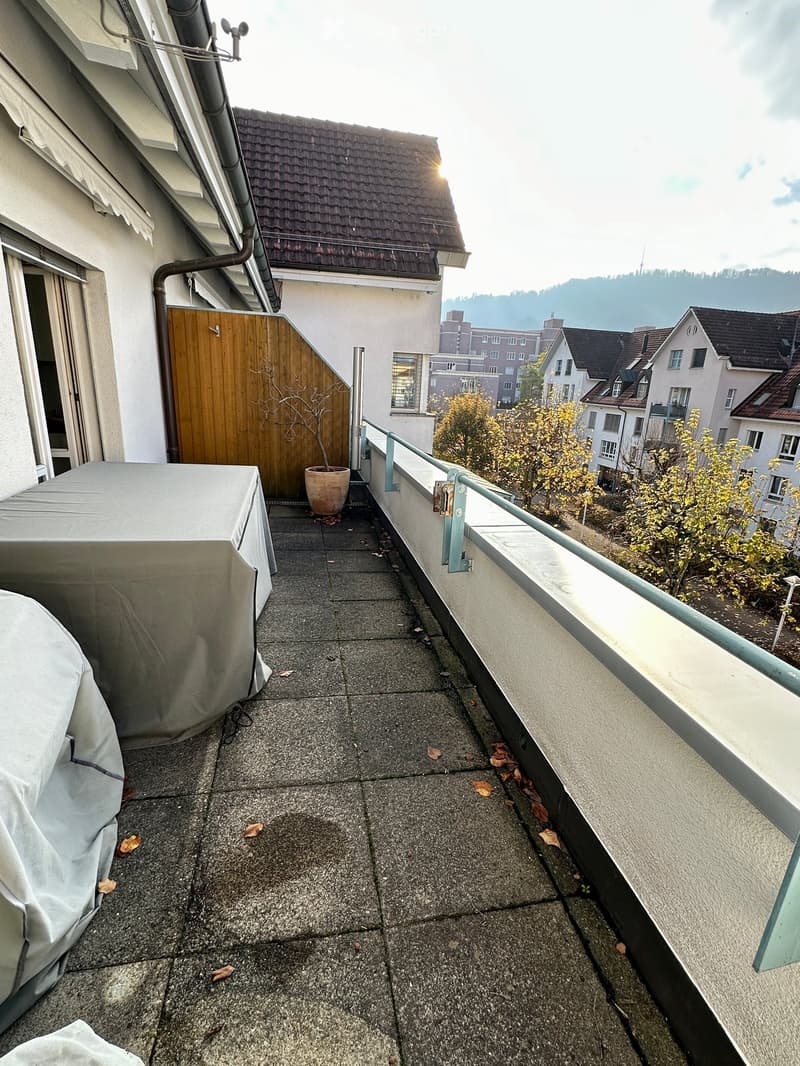 Balcony with concrete floor, covered furniture, railing, and a view of surrounding buildings and hills.