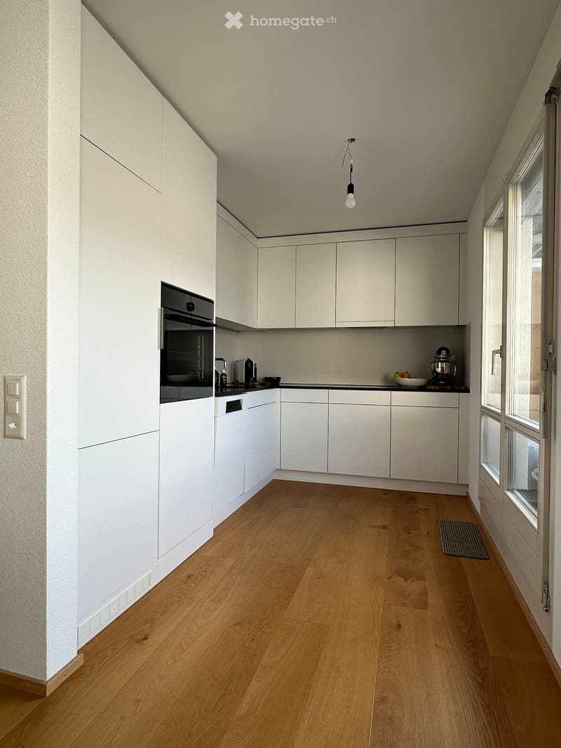 Modern kitchen with white cabinets, wooden flooring, and built-in appliances including an oven and dishwasher. A small kitchen island holds a blender and a bowl, with a window providing natural light.