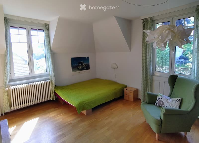Bright bedroom with wooden flooring, a single bed with green bedding, a green armchair with a decorative pillow, a radiator, a wall-mounted picture frame, and a modern hanging light fixture.