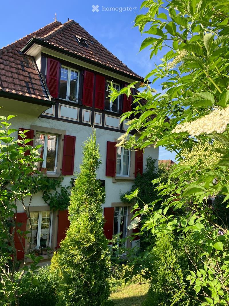 Two-story house with red shutters, white walls, and a tiled roof, surrounded by lush greenery and flowering plants.