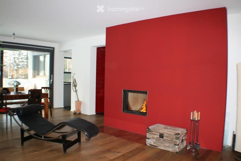 Modern living room with a red accent wall, fireplace, black leather sofa, dining table, and chairs.