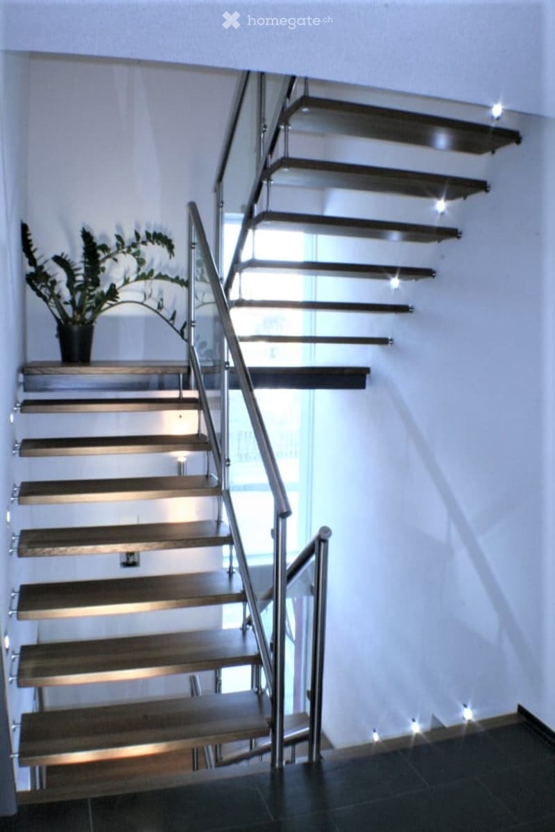 Modern staircase with wooden steps and metal railing, illuminated by small spotlights, leading to an upper level with a potted plant on the landing.