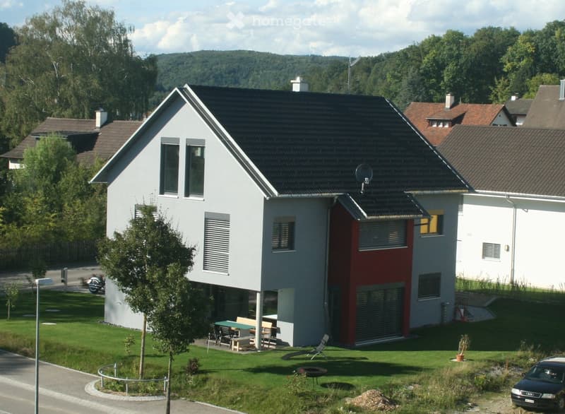 Modern two-story house with white and red exterior, large windows, and a well-maintained lawn with outdoor furniture and a barbecue area.
