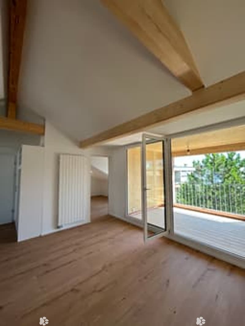 Bright, unfurnished room with wooden flooring, exposed wooden beams, large windows, and a balcony access door.