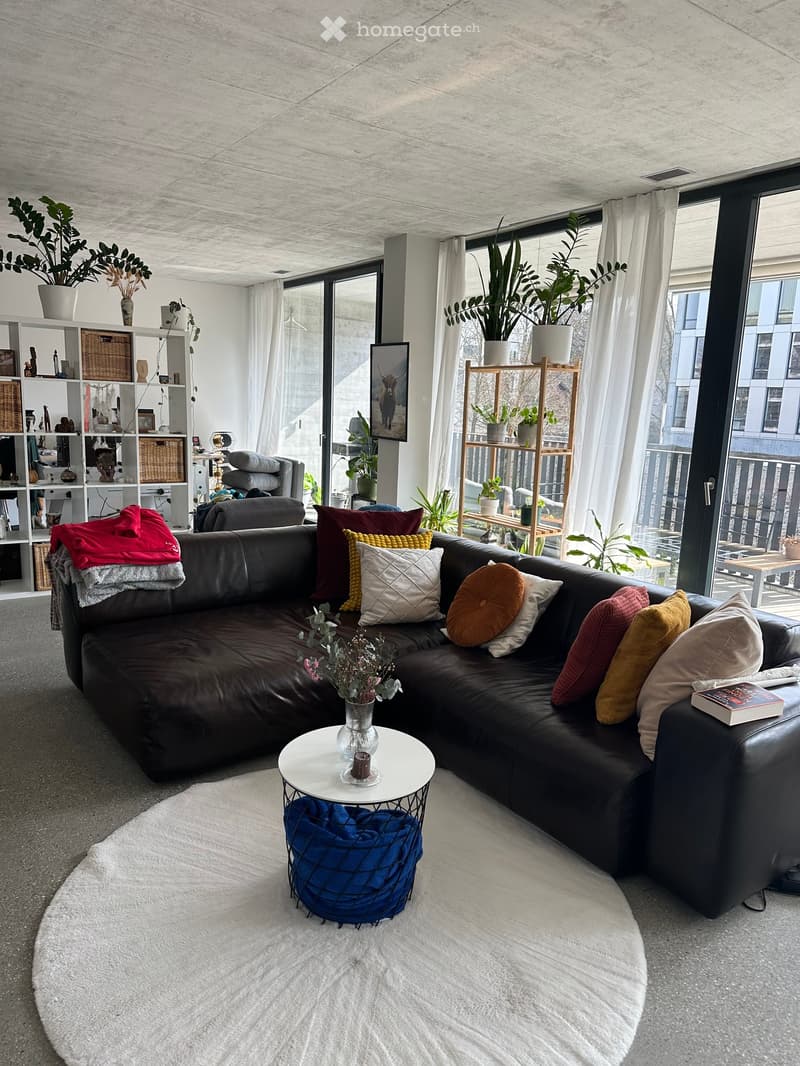 Modern living room with a large sectional sofa, colorful pillows, a white coffee table with a vase, a bookshelf, and large windows with a view.