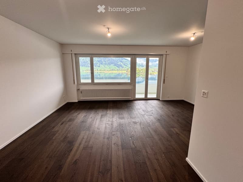 Empty room with wooden flooring, white walls, large windows with scenic mountain and lake views, and a radiator below the windows.