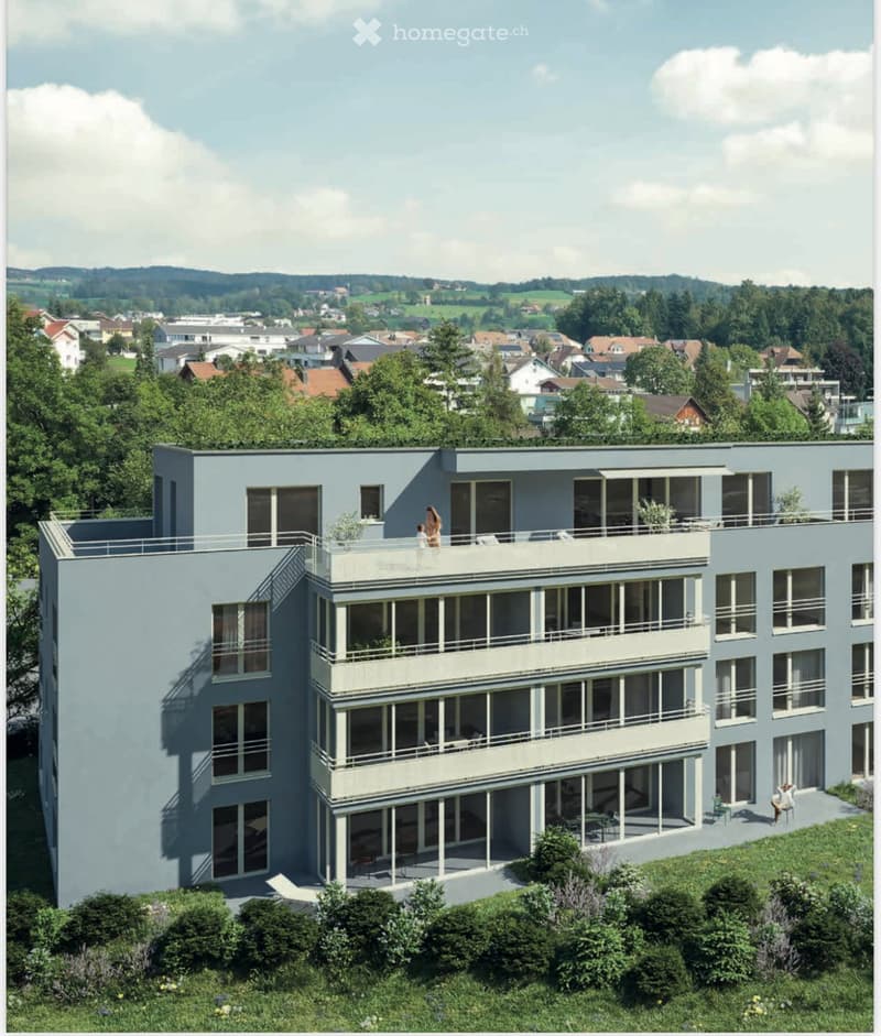 Modern, multi-story building with large windows and balconies, surrounded by greenery and situated in a suburban area with visible houses and trees in the background.