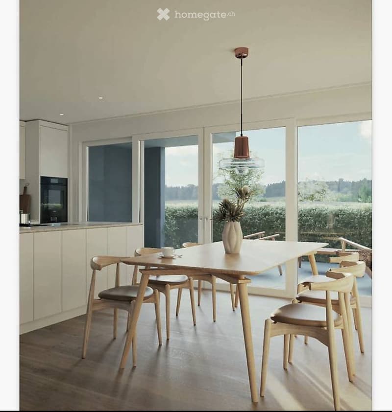Bright kitchen with a dining area, featuring a wooden dining table, six chairs, a vase with flowers, and a cup on the table. Large windows provide natural light and a view of the outdoors.
