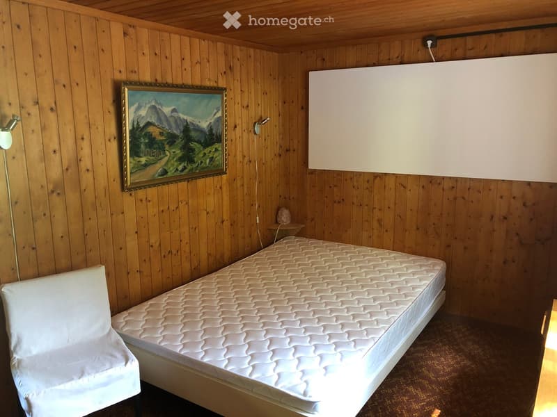 Cozy wooden-paneled room with a single bed, a white chair, a framed painting, and a large screen on the wall.