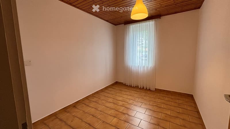 Empty room with beige walls, wooden ceiling, tiled floor, and a window with sheer curtains.