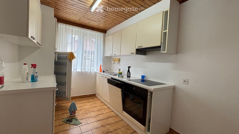A small, well-lit kitchen with white cabinets, a tiled floor, and a wooden ceiling. The kitchen features a stove, oven, sink, and various cleaning supplies on the counter.