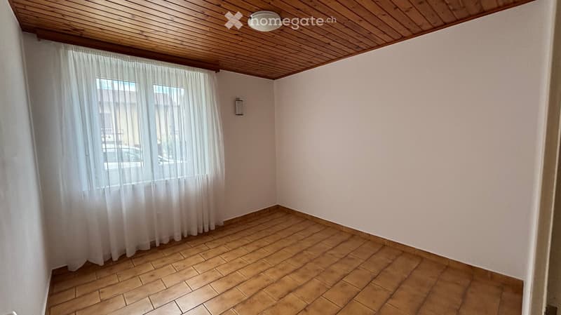 Empty room with wooden tiled floor, white walls, wooden ceiling, and large window with white curtains.