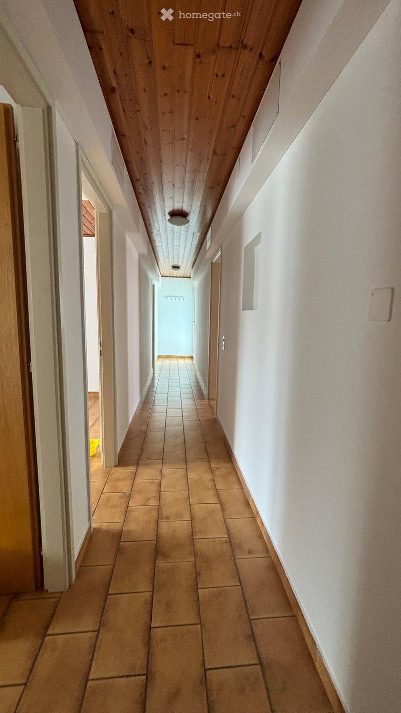 A narrow hallway with tiled flooring, white walls, and wooden ceiling beams. Doors are visible on both sides, leading to other rooms.
