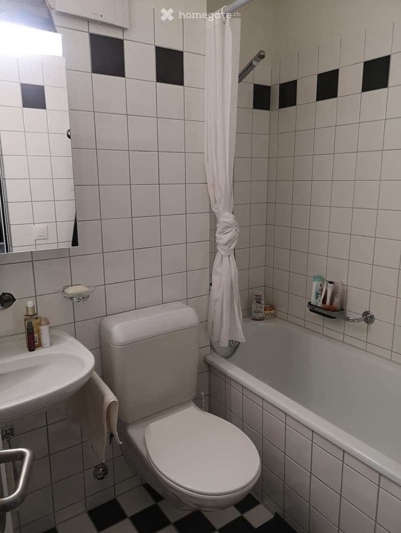 A small bathroom with white tiled walls, a toilet, a sink, a bathtub, and a mirror. A towel hangs on a rod, and various toiletries are placed on the sink and bathtub ledge.